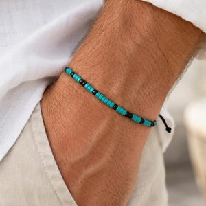 May include: A thin bracelet featuring a repeating pattern of small, flat, circular turquoise and black beads strung on a dark cord. The design consists of alternating segments of turquoise beads separated by single black beads, secured with a knot.