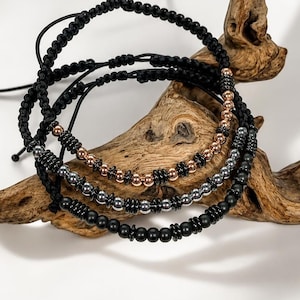 Könnte beinhalten: Drei verstellbare Armbänder aus schwarzer Kordel mit geknoteten Details. Ein Armband hat roségoldfarbene Perlen, eines metallisch silberfarbene Perlen und eines matte schwarze Perlen. Jedes Armband enthält kleine zylindrische Abstandshalter zwischen den runden Perlen.