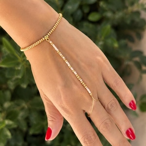 Könnte beinhalten: Ein goldfarbenes Perlenarmband, das durch eine zarte Kette aus weißen und goldenen Perlen mit einem Ring verbunden ist. Das Armband besteht aus einem durchgehenden Strang kleiner metallisch goldener Perlen, die Verbindungskette wechselt ab.