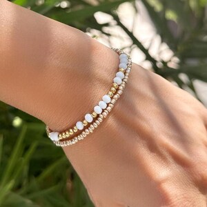 May include: Two gold beaded bracelets with white beads are worn on a wrist. The bracelets are stacked, with one featuring small gold beads and the other, white beads. Two gold rings are on the fingers.