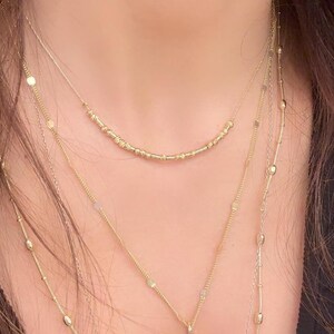 May include: A layered gold necklace set. The necklaces vary in length and design, including a beaded strand, a delicate chain with a Hamsa hand pendant, and chains with small, round and oval accents. The jewelry is worn against a dark background.