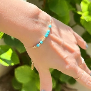 May include: A delicate gold chain bracelet with small turquoise beads. The bracelet is on a person's wrist.