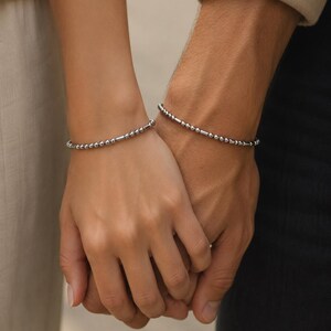 Könnte beinhalten: Nahaufnahme von zwei Handgelenken, jeweils mit einem silbernen Perlenarmband. Die Armbänder bestehen aus kleinen, runden Perlen und zylindrischen Stäben. Die Hände sind gefaltet, was auf eine Verbindung oder Beziehung hindeutet.