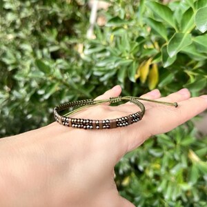 May include: A handmade bracelet with a woven design. The bracelet features small, dark beads and rectangular brown beads, strung on a green cord. The adjustable cord allows for a comfortable fit. The bracelet is displayed against a backdrop of green foliage.