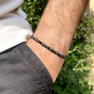 Könnte beinhalten: Ein schwarzes Perlenarmband mit minimalistischem Design. Das Armband besteht aus kleinen, dunklen Perlen, die auf einer dünnen Schnur aufgereiht sind. Das Armband wird am Handgelenk getragen, mit einem weißen Hemd und einer dunkelgrauen Hose im Hintergrund.
