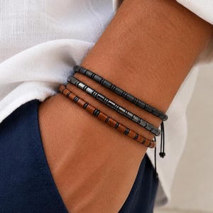 May include: A set of three stacked bracelets featuring cylindrical beads. The top bracelet is matte black, the middle is metallic gunmetal gray, and the bottom is a warm brown tone. Each bracelet is constructed with small, uniform beads.