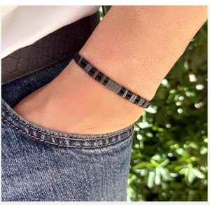 Könnte beinhalten: Nahaufnahme eines Armbands mit rechteckigen grauen und schwarzen Perlen. Das Armband wird am Handgelenk einer Person getragen, mit einer Jeans-Tasche und einem weißen Hemd im Hintergrund. Das Armband hat einen Umfang von etwa 18 cm.