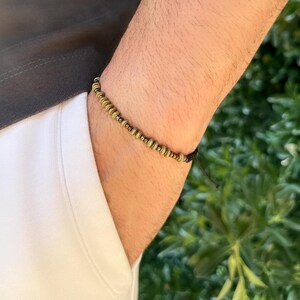 May include: A beaded bracelet with small, round, gold-colored beads strung on a black cord. The bracelet is worn on a wrist, with a white shirt and a gray shirt visible in the background. The background is a blurred green.