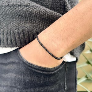 Puede incluir: Un brazalete negro de cuentas con un diseño sencillo. El brazalete está hecho de pequeñas cuentas rectangulares que están enhebradas en un cordón delgado.