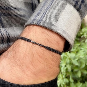 Könnte beinhalten: Ein schwarzes Perlenarmband mit einem schwarzen Band. Das Armband befindet sich an einem Handgelenk.