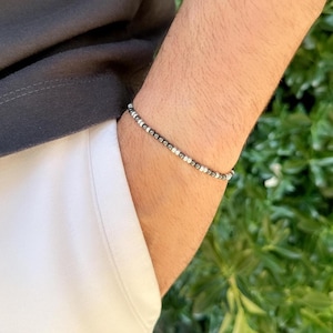 Könnte beinhalten: Ein Perlenarmband mit kleinen, runden Perlen in abwechselnd Schwarz, Silber und Weiß. Das Armband wird am Handgelenk getragen, mit einem dunkelgrauen Hemd und weißer Kleidung im Hintergrund. Das Armband hat ein schlichtes, minimalistisches Design.