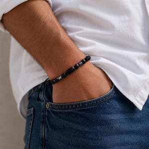 Könnte beinhalten: Ein Perlenarmband mit mattschwarzen, kugelförmigen Perlen, die mit kleinen, zylindrischen, silberfarbenen Metallelementen abwechseln. Das Armband enthält eine zentrale, texturierte schwarze Perle und wird mit einer dünnen schwarzen Kordel geschlossen.