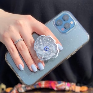 May include: A light blue phone case with a sparkly finish and a white phone grip with a blue evil eye charm.