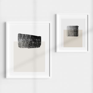 May include: Two framed abstract art prints. Each print features a textured black shape over a beige rectangle, set against a white background. The frames are white, creating a minimalist aesthetic.
