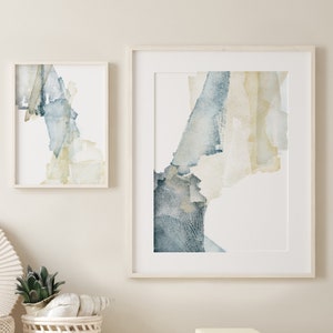 以下が含まれることがあります： 白い壁に、青と黄色の水彩画の抽象画が2つ額装されて飾られています。その下には、白い棚があり、白い花瓶、編みこまれたバスケットに入った多肉植物、そして本が積み重ねられています。