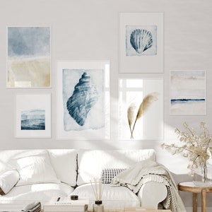 May include: A white couch with a white throw blanket and a coffee table with books and a diffuser. There are five framed watercolor paintings on the wall behind the couch. The paintings feature seashells, ocean waves, and a seashell with a brown and white feather.