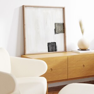 K&ouml;nnte beinhalten: Ein gerahmtes abstraktes Gem&auml;lde mit schwarzen und grauen Formen auf wei&szlig;em Hintergrund, das &uuml;ber einer Kommode aus Holz mit zwei Schubladen h&auml;ngt. Eine Vase mit getrocknetem Gras steht auf der Kommode.