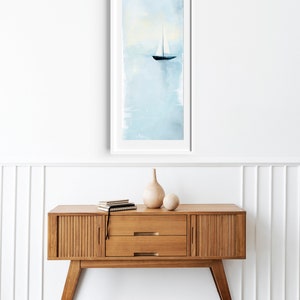 Peut inclure: Une peinture &agrave; l'aquarelle encadr&eacute;e d'un voilier sur une &eacute;tendue d'eau bleu clair. La peinture est accroch&eacute;e &agrave; un mur blanc au-dessus d'une console en bois avec deux tiroirs.