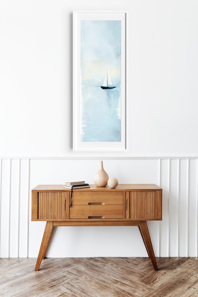 Peut inclure: Une peinture &agrave; l'aquarelle encadr&eacute;e d'un voilier sur une &eacute;tendue d'eau bleu clair. La peinture est accroch&eacute;e &agrave; un mur blanc au-dessus d'une console en bois avec deux tiroirs.