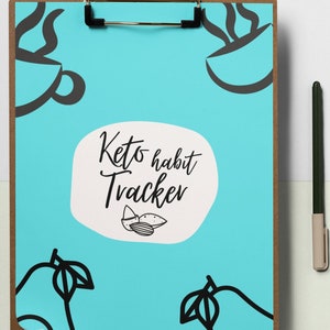 Op de afbeelding: Een blauwe clipboard met een bruin frame en een metalen clip. De clipboard heeft een witte cirkel met de tekst "Keto habit Tracker" en een tekening van amandelen. Er zijn tekeningen van avocado's en koffiekopjes op de clipboard.