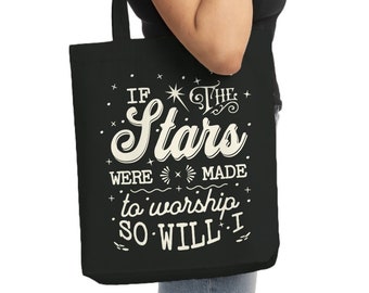May include: Black tote bag with white text that reads "If the stars were made to worship so will I." The bag is held by a person wearing a black shirt and blue jeans. The tote bag has a simple design with a rectangular shape and long handles.