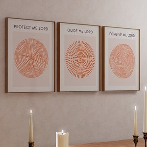 May include: Three framed prints with abstract geometric designs in orange on a light pink background. The prints have the text "Protect Me Lord", "Guide Me Lord", and "Forgive Me Lord" in black. The prints are hanging on a light beige wall above a wooden table with three white candles.