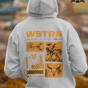 May include: A gray hooded sweatshirt with a graphic print on the back. The graphic features the word "WSTRN" in large letters, followed by the words "GRILL AND ROOTS". Below the text are four squares, each containing a different image. The images include a scorpion, a cowboy riding a horse, a bird, and a skull. The graphic is in a vintage style, with a distressed look.