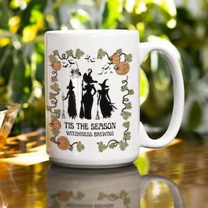 Puede incluir: Taza de cerámica blanca con un diseño negro y verde con tres brujas y el texto "Tis the season Witchiness Brewing".