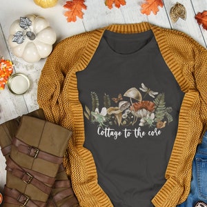 May include: A flat lay featuring a dark gray t-shirt with a mushroom and foliage graphic, the text "Cottage to the core", a mustard yellow cardigan, brown boots, blue jeans, and fall-themed decorations.