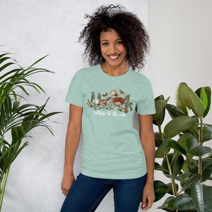 May include: A light green t-shirt featuring a watercolor illustration of mushrooms, plants, and insects. The text "Cottage to the core" is written in a cursive font below the illustration. The shirt is made of a soft material.