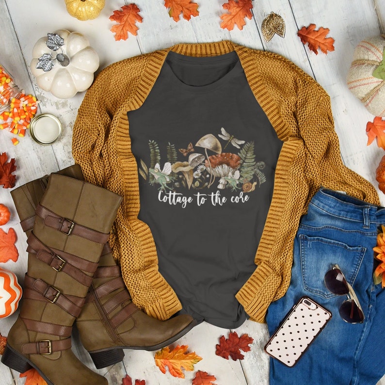 May include: A flat lay featuring a dark gray t-shirt with a mushroom and foliage graphic, the text "Cottage to the core", a mustard yellow cardigan, brown boots, blue jeans, and fall-themed decorations.
