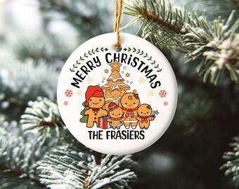May include: White round Christmas ornament with a gingerbread family standing in front of a gingerbread tree. The ornament says "Merry Christmas The Frasiers" in black text.