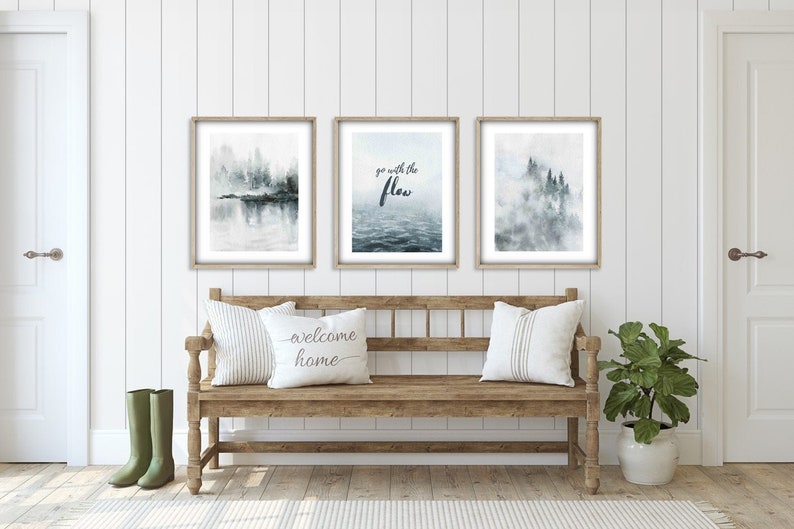 Op de afbeelding: Drie ingelijste prints met een aquarelontwerp. De prints tonen een mistige bos sc&egrave;ne, een citaat dat zegt "go with the flow", en een andere mistige bos sc&egrave;ne. De prints hangen aan een witte muur boven een houten bank met kussens. Een plant in een pot staat op de vloer rechts van de bank.