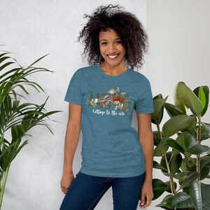 May include: Teal short-sleeve t-shirt with a nature-themed graphic. The graphic features mushrooms, plants, and butterflies, with the text "Cottage to the core" in white script. The model is wearing blue jeans.