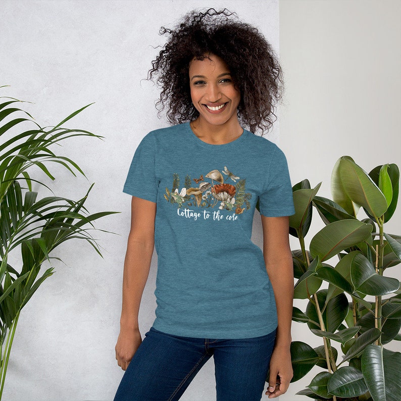 May include: Teal short-sleeve t-shirt with a nature-themed graphic. The graphic features mushrooms, plants, and butterflies, with the text "Cottage to the core" in white script. The model is wearing blue jeans.