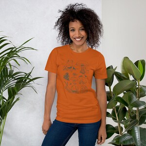 K&ouml;nnte beinhalten: Eine Frau tr&auml;gt ein orangefarbenes T-Shirt mit einer Schwarzwei&szlig;illustration von Pilzen, einer Schnecke, einem Frosch und einer Kiefer.