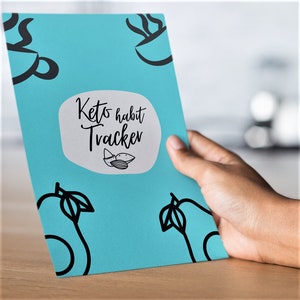 Op de afbeelding: Een blauw keto-gewoontetracker-journaal met een witte cirkel met de tekst "Keto habit Tracker" en een tekening van amandelen. Het journaal heeft aan elke kant een tekening van een avocado.