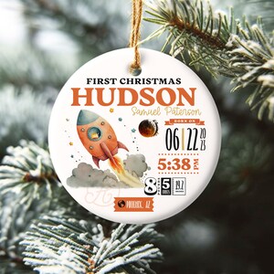 May include: White ceramic Christmas ornament with a rocket ship, stars, and planets. The ornament reads "First Christmas Hudson Samuel Paterson Born on 06.12.20 5:38 PM 8 lbs 5 oz Phoenix, AZ"