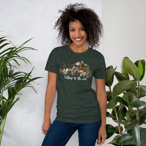 May include: Dark green t-shirt with a nature-themed graphic featuring mushrooms, ferns, and butterflies. The text "Cottage to the core" is printed in white script across the bottom of the design. The shirt is worn by a person with dark curly hair.