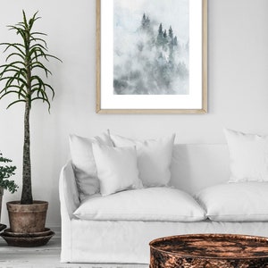 Op de afbeelding: Een ingelijst aquarel schilderij van een mistige bossc&egrave;ne met hoge, donkergroene bomen. Het schilderij is in een licht houten lijst en hangt aan een witte muur.