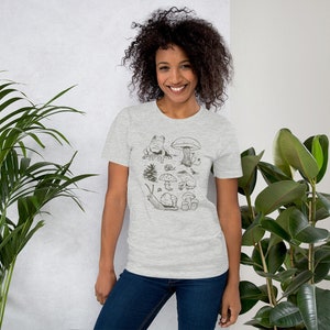 K&ouml;nnte beinhalten: Ein heidefarbenes T-Shirt mit einer Schwarzwei&szlig;illustration eines Pilzes, einer Schnecke, eines Frosches und eines Baumes. Die Illustration ist von kleineren Pilzen umgeben.