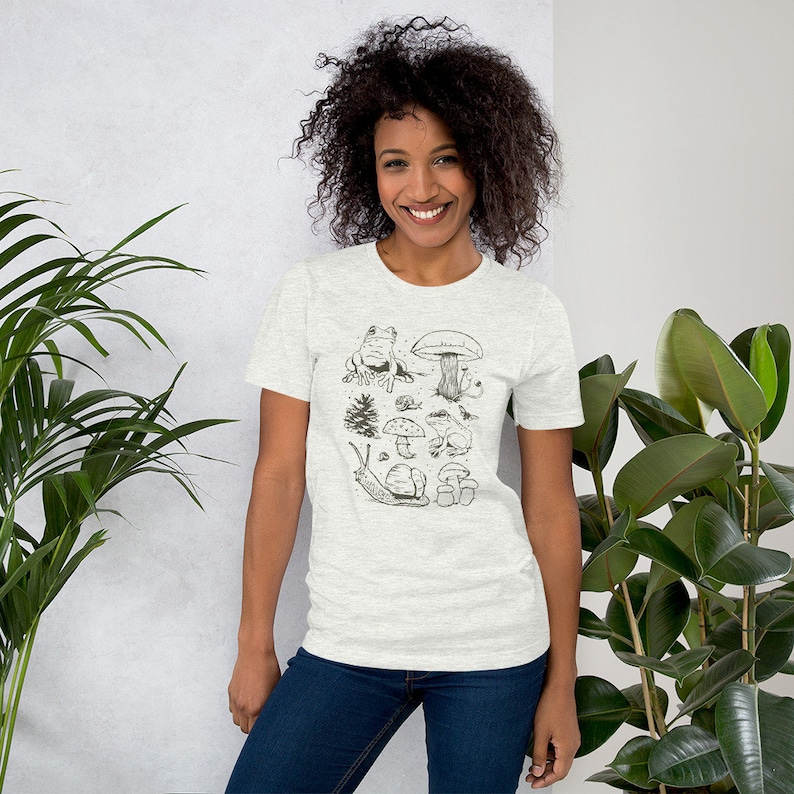 K&ouml;nnte beinhalten: Ein hellgraues T-Shirt mit einer Schwarzwei&szlig;illustration von Pilzen, einer Schnecke, einem Frosch und einem Baum.