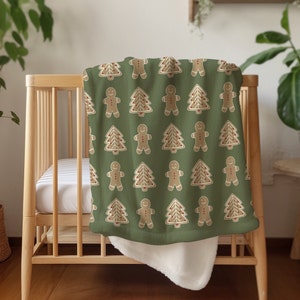 Pode incluir: Um cobertor de bebê verde com um padrão repetido de homens de gengibre e árvores de Natal. O cobertor está dobrado e drapeado sobre um berço de madeira.
