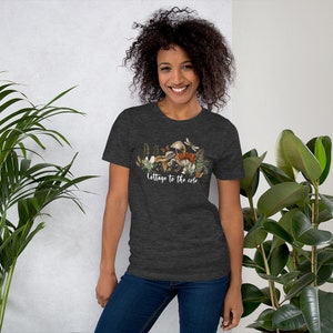 May include: Dark gray t-shirt featuring a nature-themed graphic with mushrooms, ferns, and butterflies. The text "Cottage to the core" is printed in a cursive font. The shirt is made of a soft material.