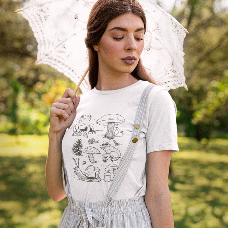 K&ouml;nnte beinhalten: Wei&szlig;es T-Shirt mit einer schwarz-wei&szlig;en Illustration von Pilzen, Fr&ouml;schen und einer Schnecke. Die Illustration ist mittig auf dem Shirt platziert.
