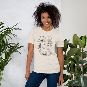K&ouml;nnte beinhalten: Ein beiges T-Shirt mit einer schwarz-wei&szlig;en Illustration eines Frosches, Pilzen, einer Schnecke und eines Baumes. Die Illustration ist mittig auf dem T-Shirt platziert.