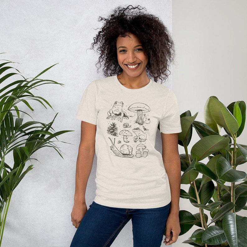 K&ouml;nnte beinhalten: Ein beiges T-Shirt mit einer schwarz-wei&szlig;en Illustration eines Frosches, Pilzen, einer Schnecke und eines Baumes. Die Illustration ist mittig auf dem T-Shirt platziert.