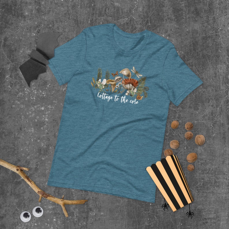 May include: Teal short-sleeve t-shirt with a watercolor illustration of mushrooms, ferns, and a dragonfly. The text "Cottage to the core" is printed below the illustration. The shirt is made of a soft material.