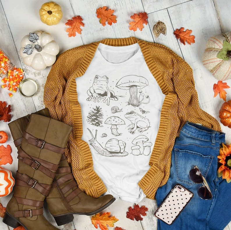 K&ouml;nnte beinhalten: Wei&szlig;es T-Shirt mit einer schwarz-wei&szlig;en Illustration einer Schnecke, Pilzen und Fr&ouml;schen. Das Shirt liegt auf einer wei&szlig;en Holzoberfl&auml;che mit einem braunen Cardigan, einer blauen Jeans und Herbstdekoration.