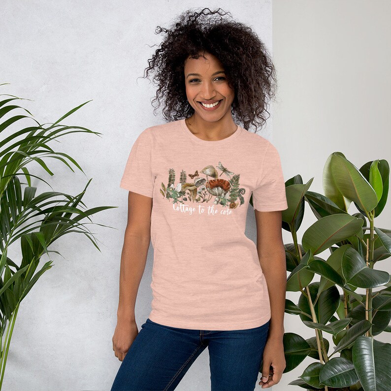 May include: A light pink t-shirt featuring a nature-themed graphic with mushrooms, plants, and insects. The text "cottage to the core" is printed in white script across the bottom of the design. The shirt is made of a soft material.